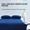 A dark blue cooling ice-silk fitted sheet with two pillows and a white lamp on a white surface.