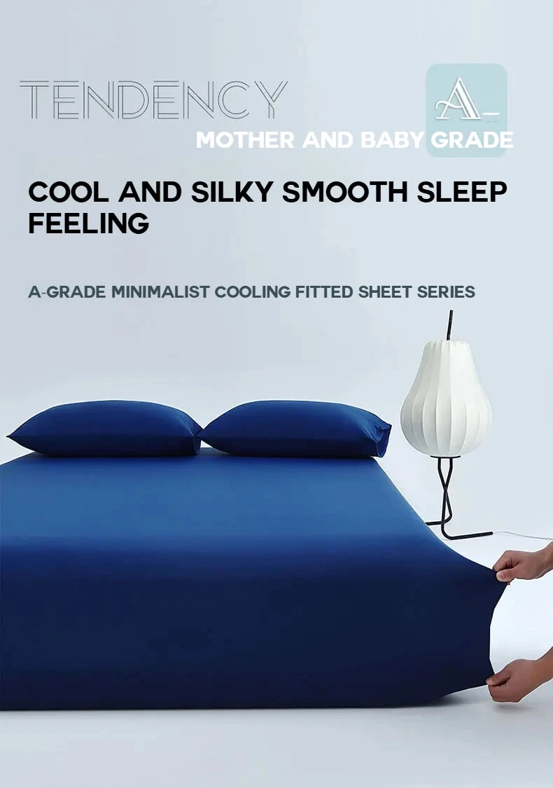 A dark blue cooling ice-silk fitted sheet with two pillows and a white lamp on a white surface.