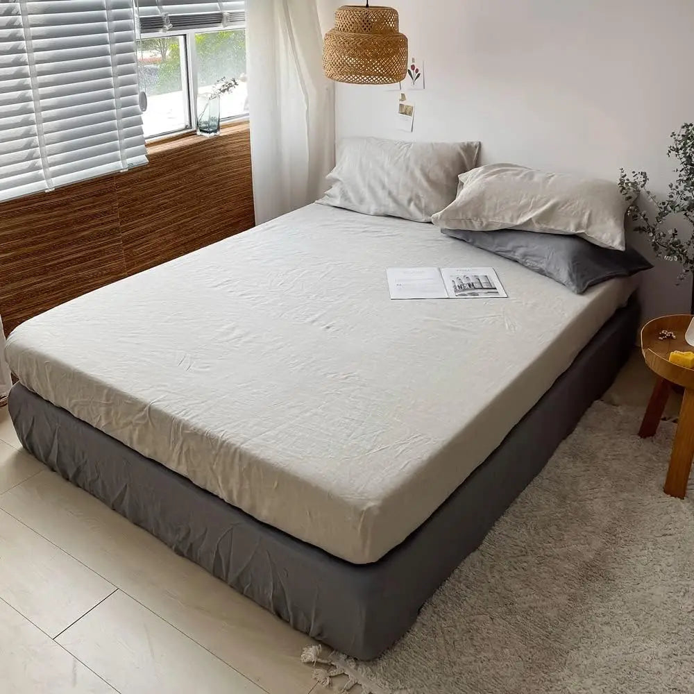 A white linen fitted sheet is laid on a bed with gray bedding and pillows in a bright room.