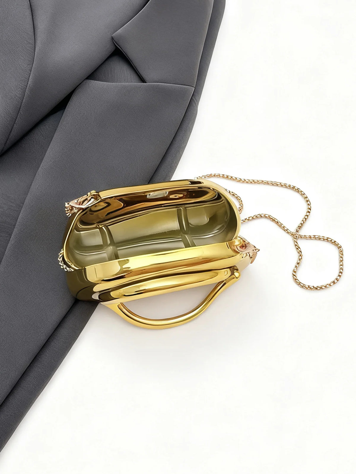 A small, rectangular gold metal clutch bag with a gold chain strap is shown against a gray fabric background.