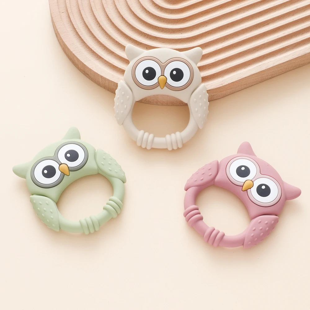 A set of owl-shaped silicone teethers and rattles in white, light green, and pink colors resting on a wooden surface.