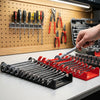 A hand is placing a wrench into a red organizer tray filled with various tools and pliers on a white surface.