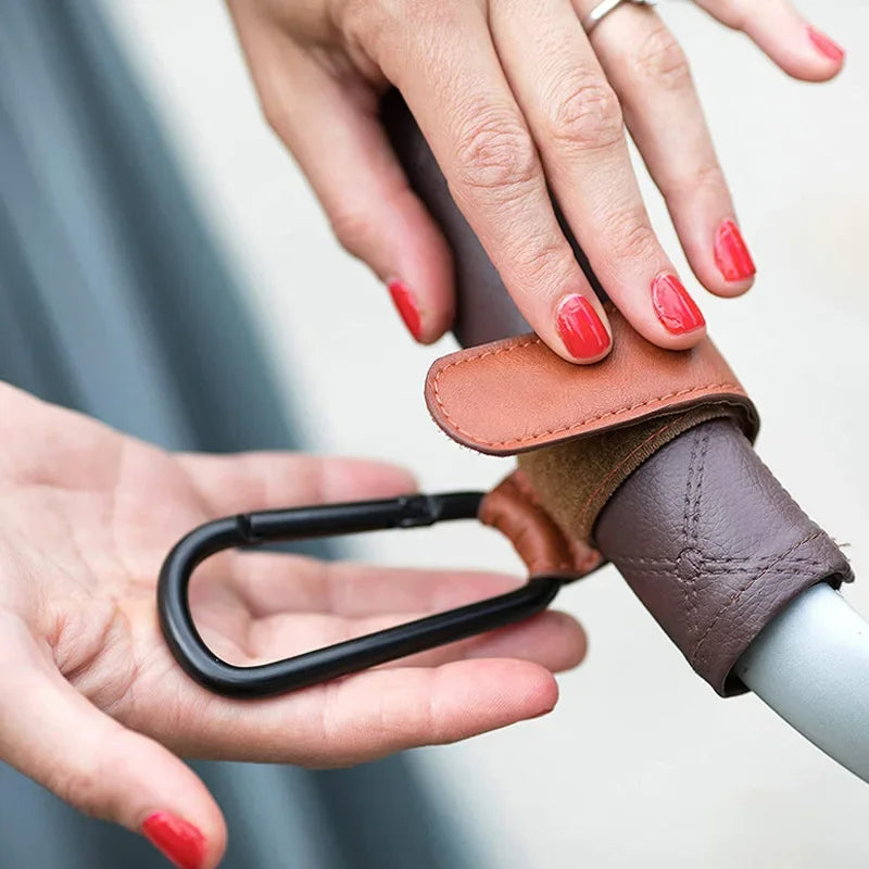 A hand is holding a brown and gray stroller hook with a PU leather baby bag organizer.