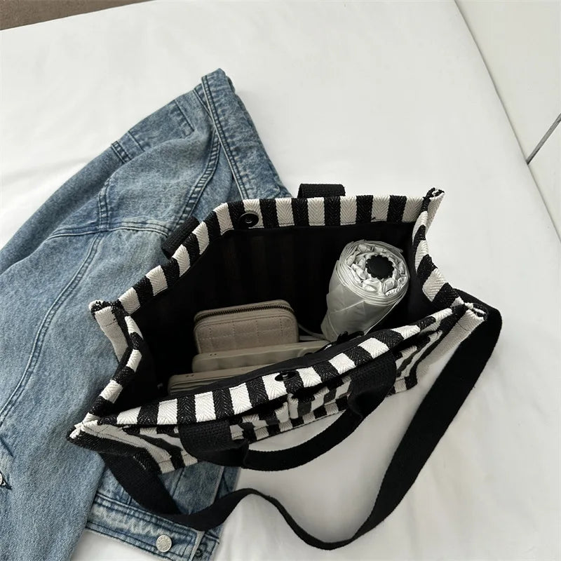 A black and white striped canvas tote bag with inner pockets resting on a white surface next to denim jeans.