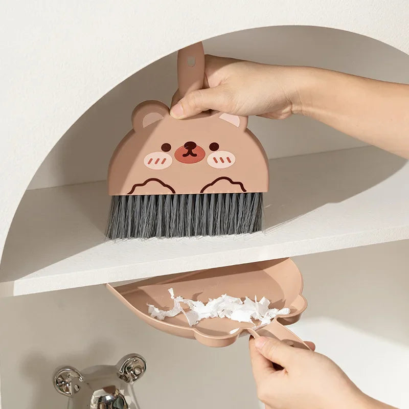 A hand is holding a small, brown cartoon bear broom and dustpan set against a white shelf.