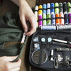 A hand is shown holding a compact portable sewing kit with various threads and tools inside a black pouch.