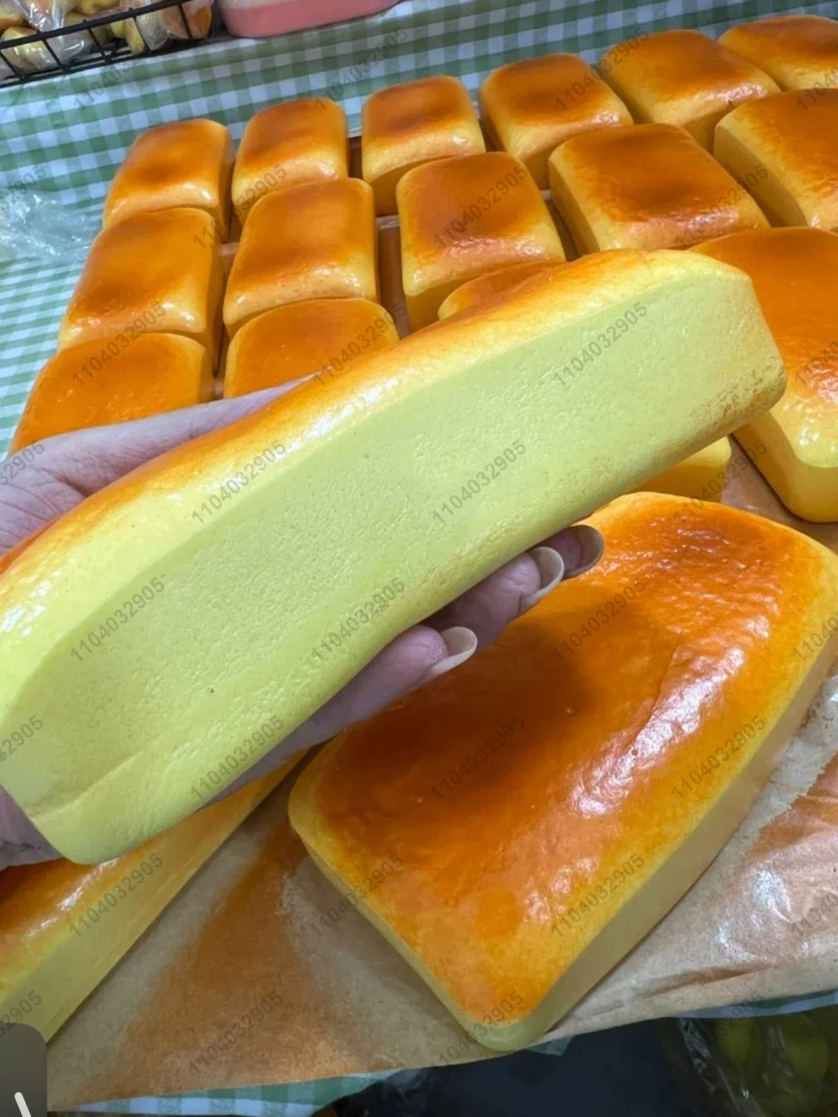 A hand holds a yellow slow-rising grilled rice cake squishy toy next to a tray of similar baked goods.