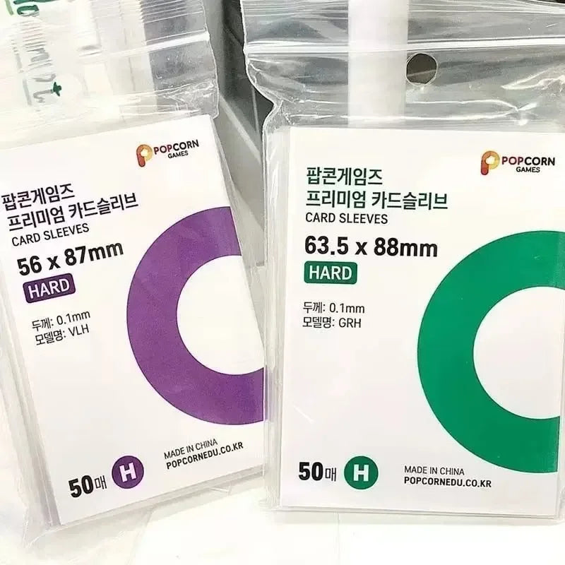 Deux paquets de pochettes pour cartes POPCORN GAMES, une violette et une verte, montrant les dimensions et la quantité.