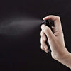 Una mano sostiene un atomizador de perfume negro, rociando una fina niebla sobre una superficie oscura.