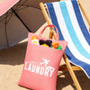 A pink laundry bag is placed next to a wooden deck chair with a striped towel and an open umbrella on a sandy surface.