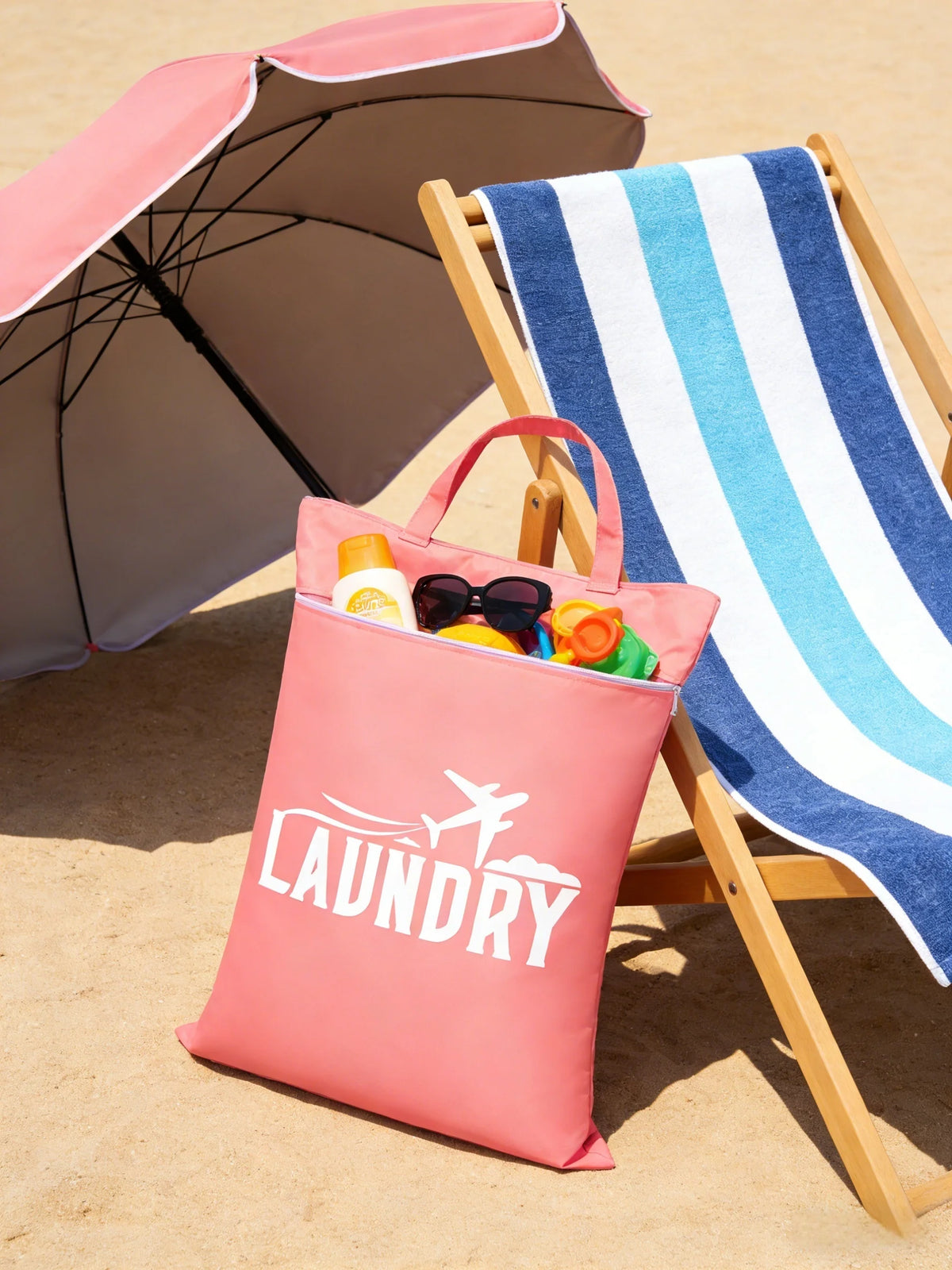 A pink laundry bag is placed next to a wooden deck chair with a striped towel and an open umbrella on a sandy surface.
