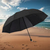 A black oversized folding umbrella is open on a sandy beach with ocean waves in the background.