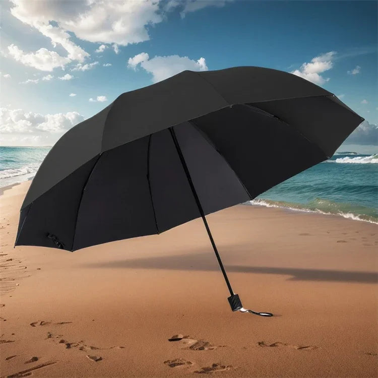 A black oversized folding umbrella is open on a sandy beach with ocean waves in the background.