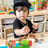 A toddler wearing a black hat is playing with colorful magnetic building blocks on a wooden table.