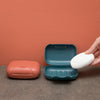 A hand opens a teal and white leak-proof travel soap box on a brown surface against a terracotta wall.