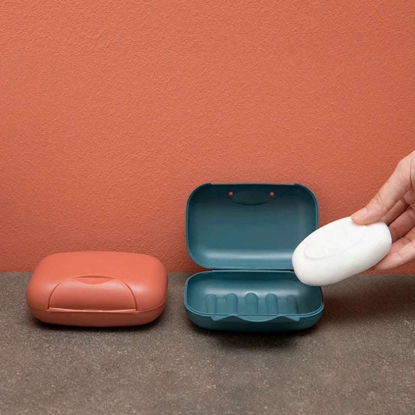 A hand opens a teal and white leak-proof travel soap box on a brown surface against a terracotta wall.