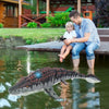 A man and a young girl are sitting on a wooden dock near water, with a Mosasaurus toy floating in the water.