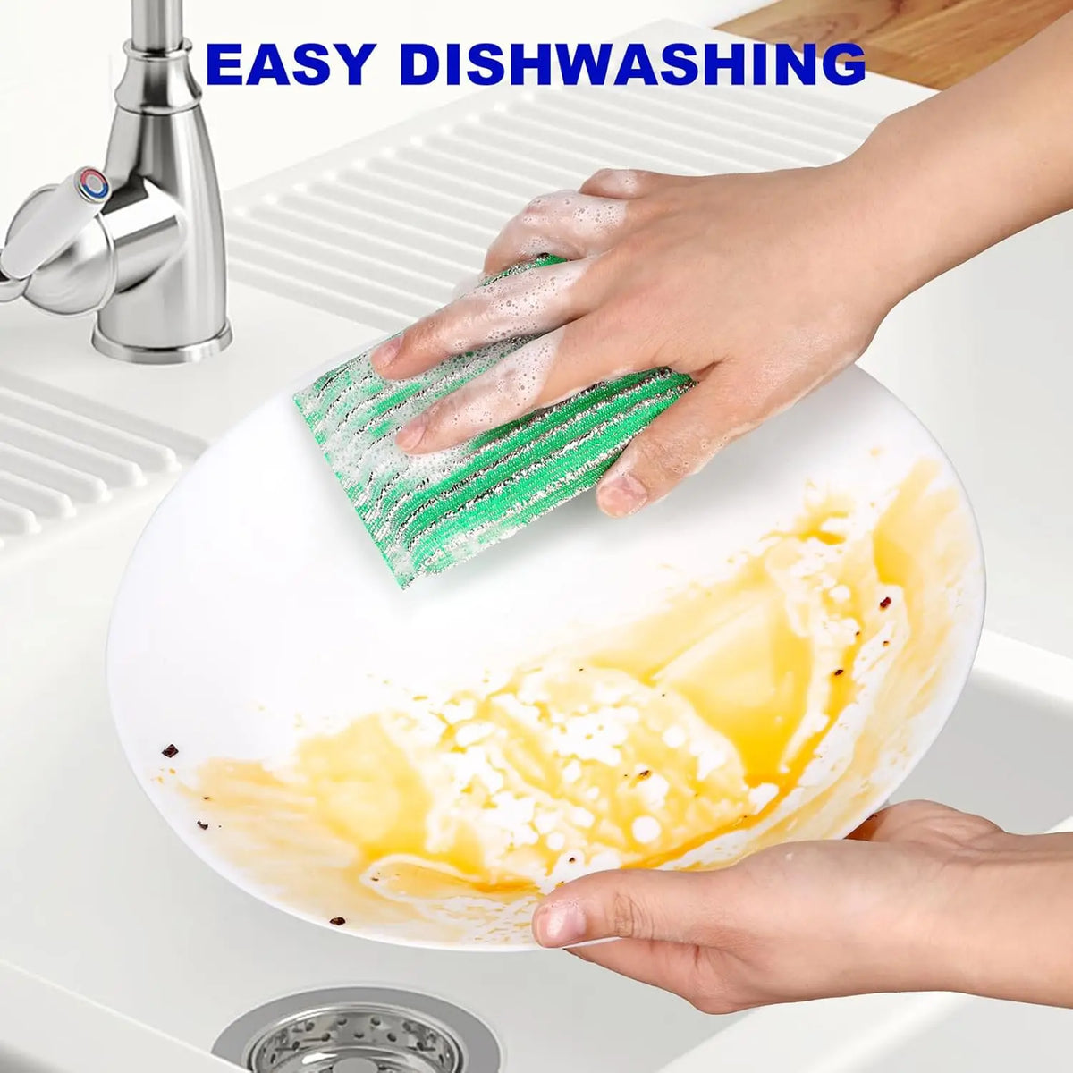 A hand is using a green scouring sponge to wash a plate in a kitchen sink.
