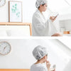 A woman wearing a white shower cap is looking at her phone in a bright bathroom setting.