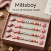 A box of personalized baby pacifier clips featuring names like Emma, Olivia, and Sophia, displayed on a wooden surface.