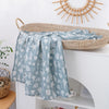 A light blue muslin baby swaddle blanket with a white leaf pattern draped over a woven basket on a white shelf.