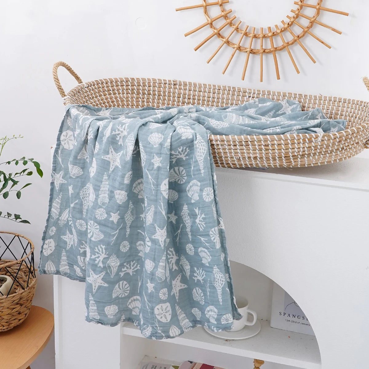 A light blue muslin baby swaddle blanket with a white leaf pattern draped over a woven basket on a white shelf.