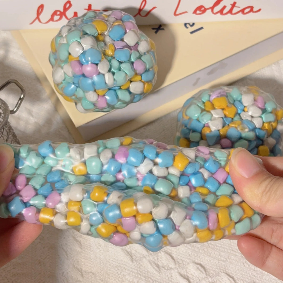 A hand holds a squishy sensory stress ball filled with multicolored soft plastic beads.