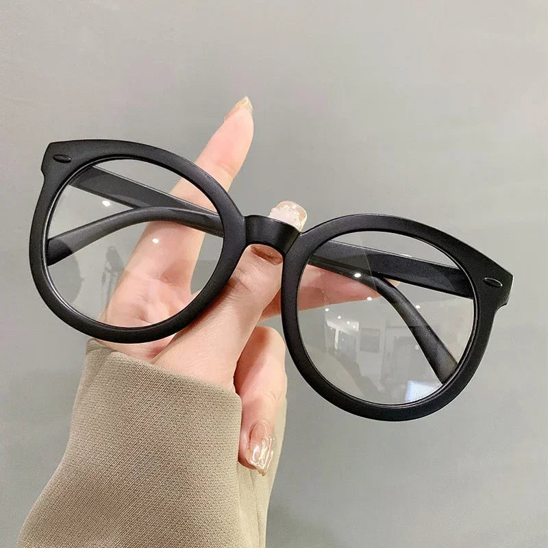 Eine Hand hält eine schwarze, überdimensionierte blaue Lesebrille vor einem hellgrauen Hintergrund.