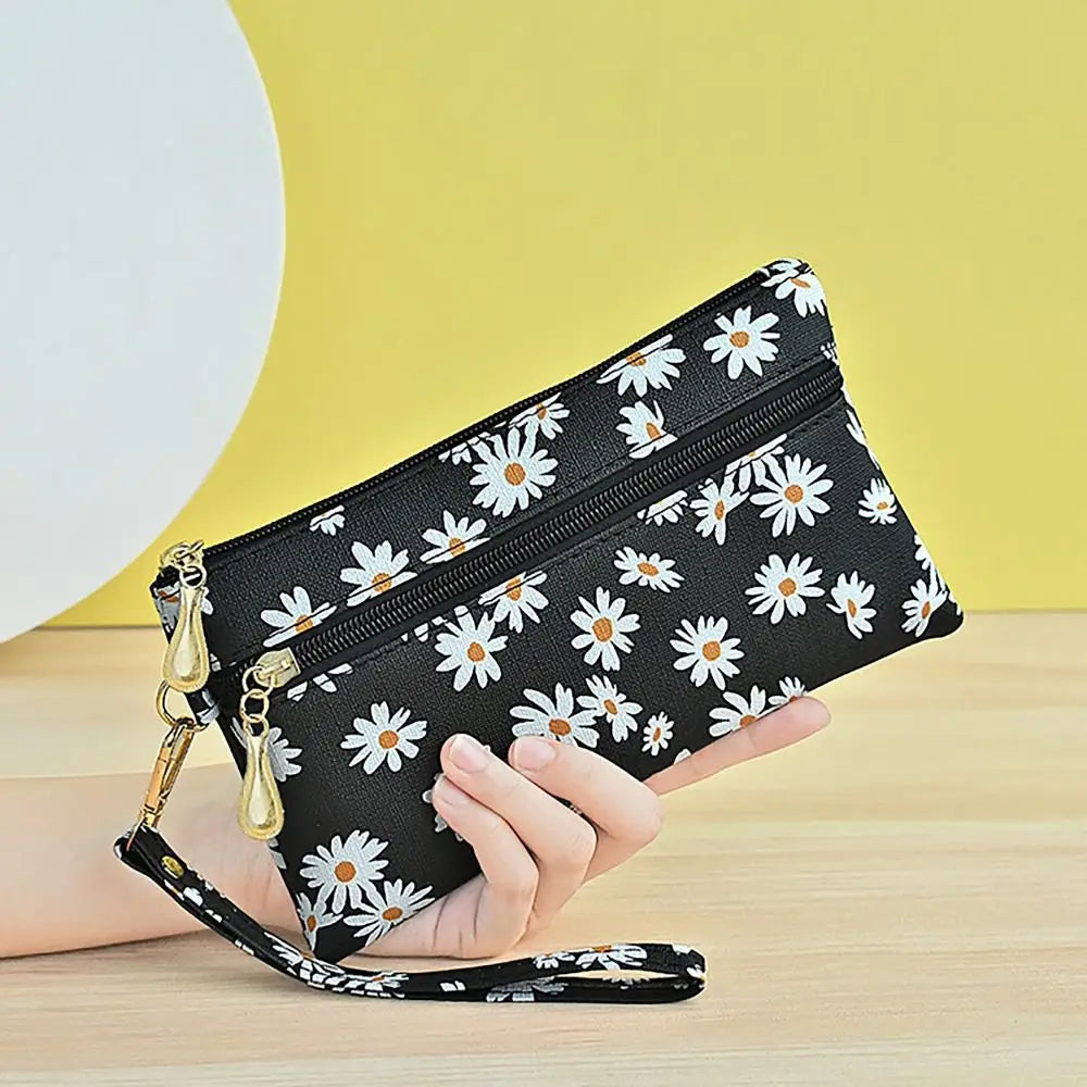 A black faux leather wristlet wallet with a daisy print pattern and gold hardware is held by a hand against a yellow backgrou