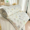 A white quilt with a pastoral print featuring yellow and green floral designs is rolled up on a white surface.