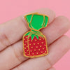 A hand holds a green and red enamel pin shaped like a strawberry with a green leafy top.