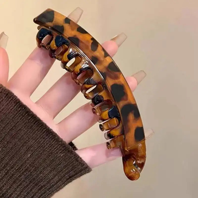 A hand holds a tortoiseshell banana hair clip with a glossy, mottled pattern.