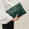 A person holds a dark green crocodile-embossed PU leather envelope clutch against a light background.