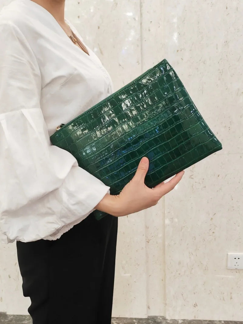 A person holds a dark green crocodile-embossed PU leather envelope clutch against a light background.