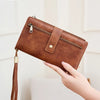 A brown PU leather long wallet with a zipper and leather strap is being held by a hand.