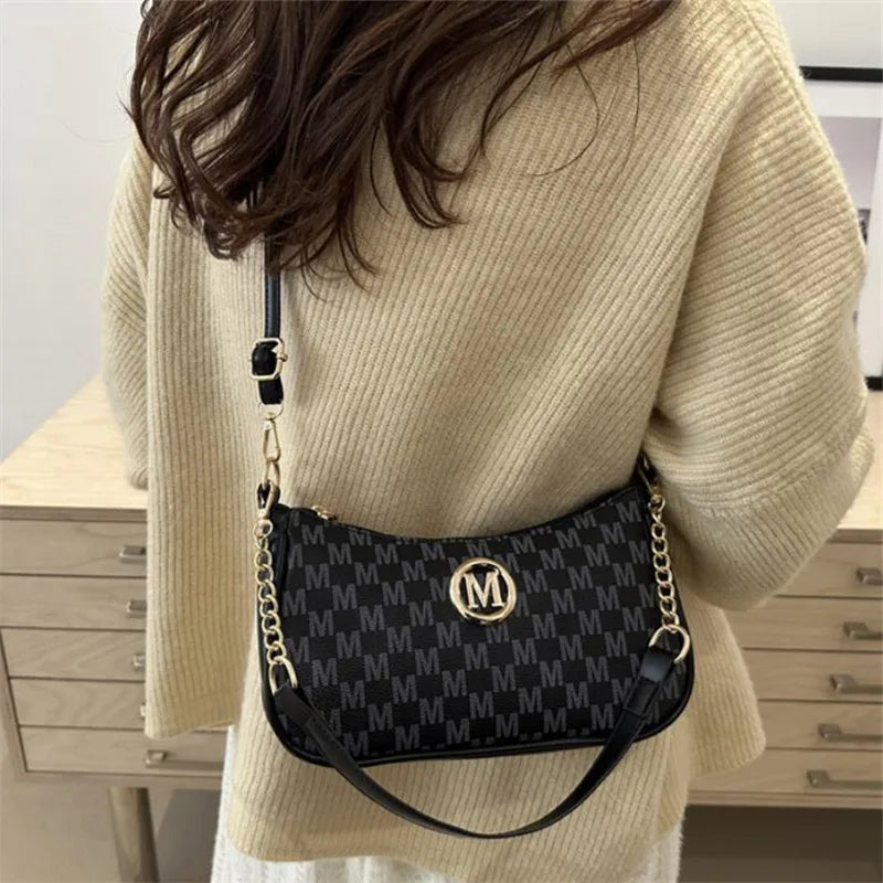 A beige sweater sleeve shows a black leather crossbody bag with a gold metal M accent.