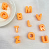 Perles en acrylique orange formant les lettres ROMV L ZOUR, éparpillées sur une surface blanche à côté d'un petit bol