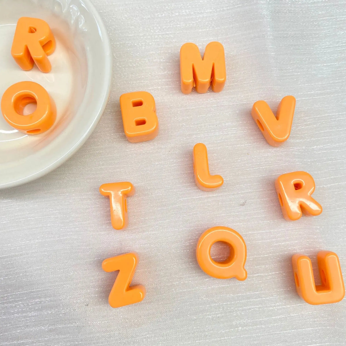 Perles en acrylique orange formant les lettres ROMV L ZOUR, éparpillées sur une surface blanche à côté d'un petit bol