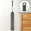 A gray microfiber duster with a handle is shown hanging on a tile, next to a wooden drawer.