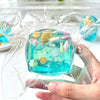 A hand holds a clear plastic bag containing a bright blue squishy ocean shell cube fidget toy.