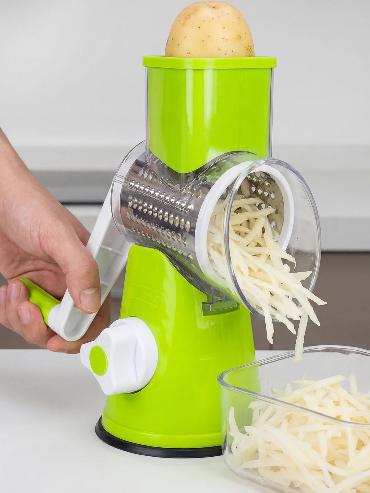 A hand is operating a lime green multi-function rotary cheese grater with a round mandoline slicer, grating a potato.