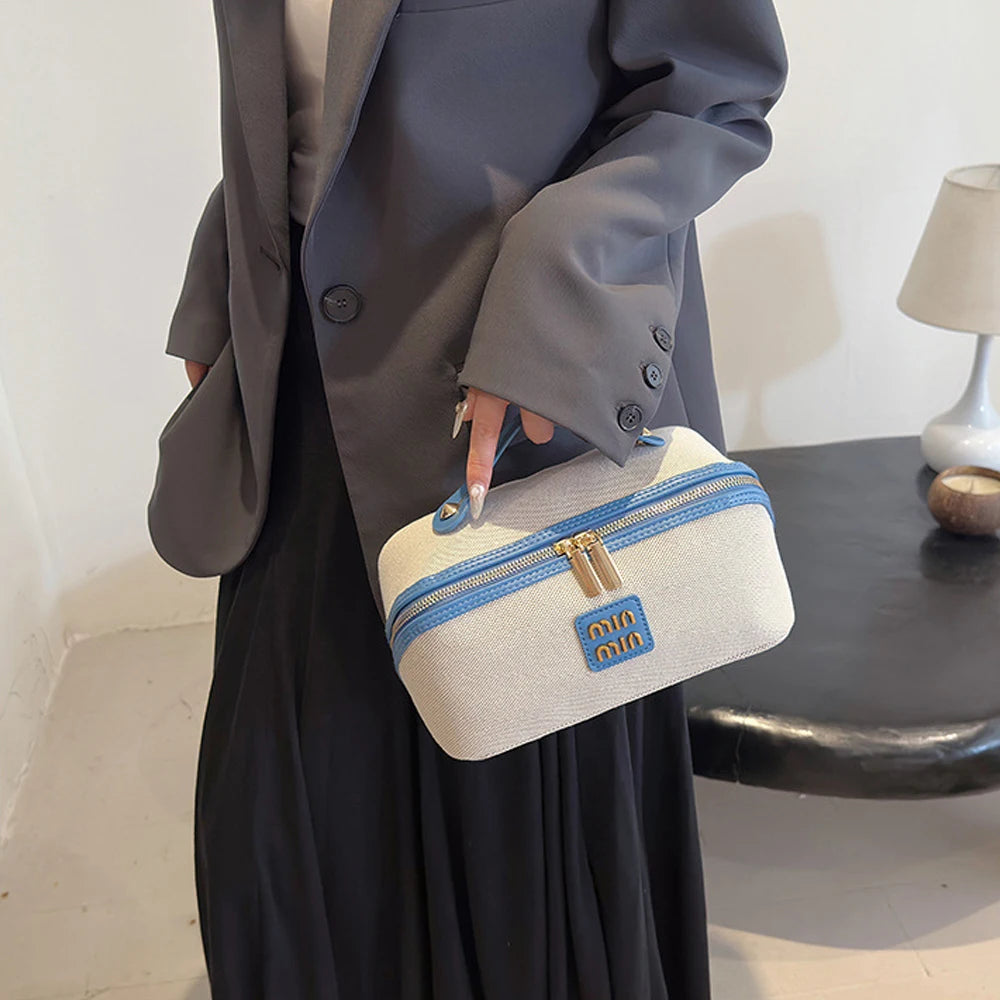 Eine Frau hält eine hellbeige Reise-Make-up-Organizer-Tasche mit blauen Akzenten und goldenen Beschlägen.