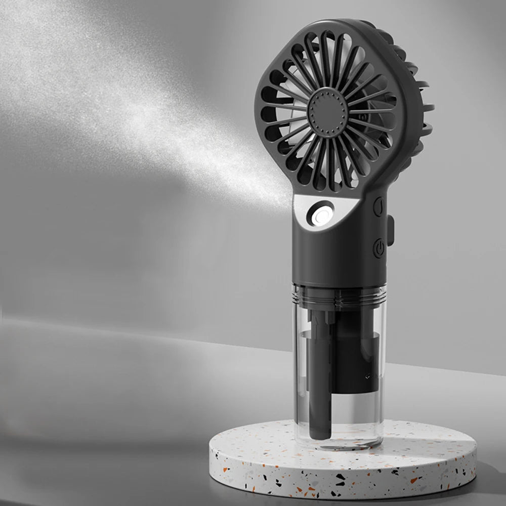 A black portable USB rechargeable mist fan with a circular grille and a clear glass container sits on a speckled white base.