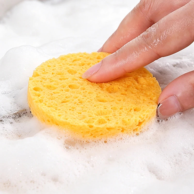 A hand is pressing a bright yellow compressed natural sponge into a pile of white soap foam.