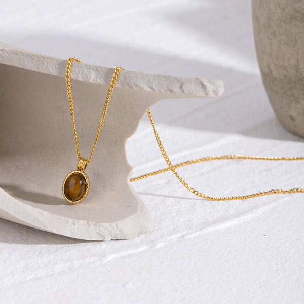 A gold chain necklace with an oval tiger's eye pendant resting on a light stone surface.