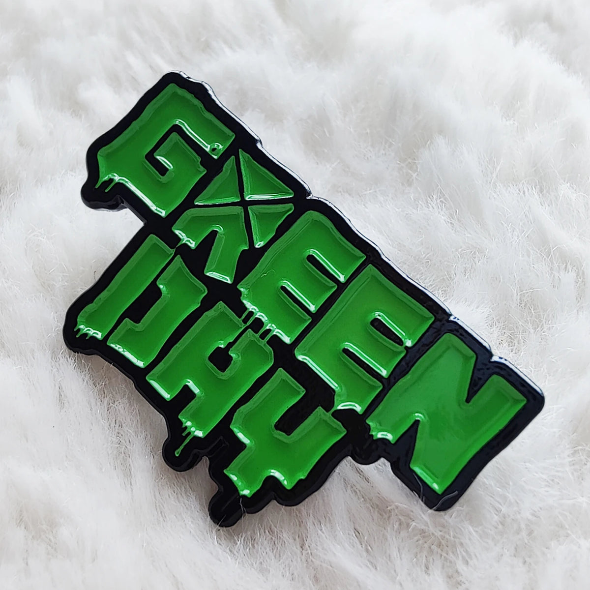 A green enamel pin shaped like the word GREEN on a white fluffy surface.