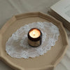 A dark jar candle sits on a white lace embroidered placemat which rests on a beige ceramic plate.
