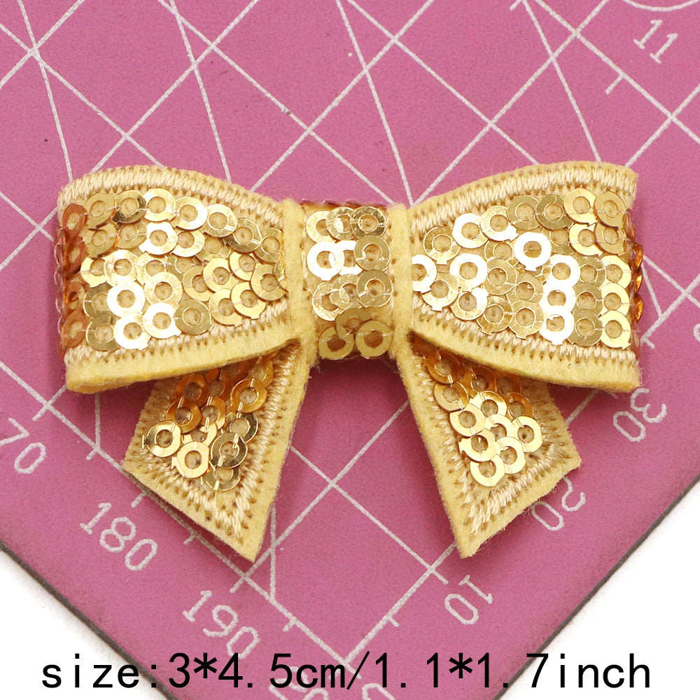 A set of five gold and bronze sequin bow patches for DIY crafts displayed on a pink grid background.