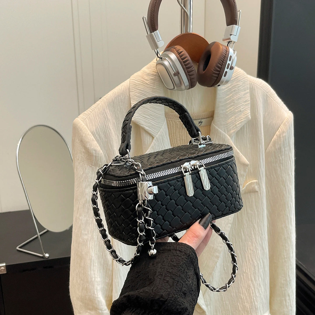 A black woven PU leather crossbody bag is displayed against a light beige shirt with headphones in the background.