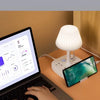 A laptop is open on a desk next to a white desk lamp and a smartphone charging on the surface.